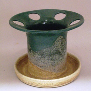 May include: A green ceramic planter with a wide, flat top and four holes. The planter has a brown and white speckled glaze and sits on a matching saucer.