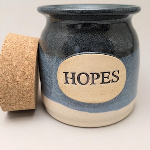 May include: A ceramic jar with a cork lid. The jar is a mix of blue and cream colors, with the word "HOPES" on a tan oval. The jar has a wide opening and a rounded shape, suitable for storage.