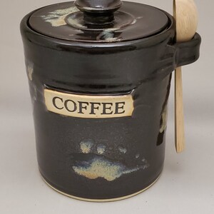 May include: Black ceramic coffee canister with a lid and a wooden spoon. The canister has a white label that says "COFFEE" and has a brown and white splatter design.