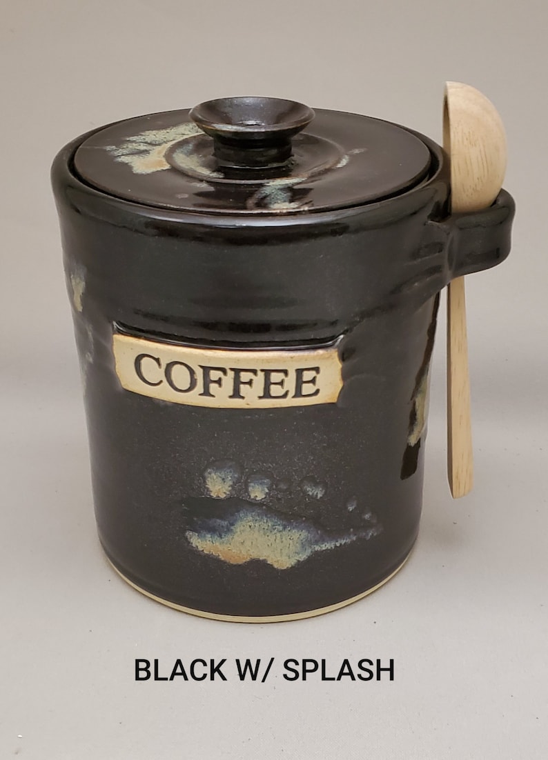 May include: Black ceramic coffee canister with a lid and a wooden spoon. The canister has a white label that says "COFFEE" and has a brown and white splatter design.