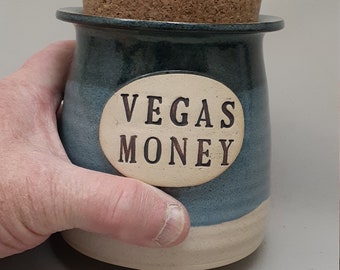 May include: A blue and white ceramic jar with a cork lid. The jar has a white oval label with the text "Vegas Money" in black lettering.