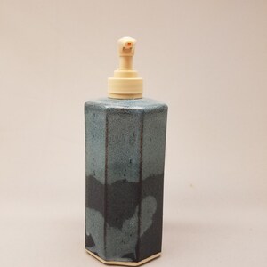 May include: A hexagonal ceramic soap dispenser featuring a two-tone glaze with a speckled light blue top section and a solid dark charcoal bottom section. The vessel is topped with a functional, light beige plastic pump mechanism for dispensing.