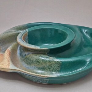 May include: A ceramic serving dish with a central circular bowl surrounded by a curved, asymmetrical outer section. The piece features a glossy deep green glaze, with areas of exposed tan clay and mottled blue-green transitions along the edges.