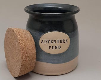 May include: A blue and white ceramic jar with a cork lid. The jar has a white oval label that says "Adventure Fund".
