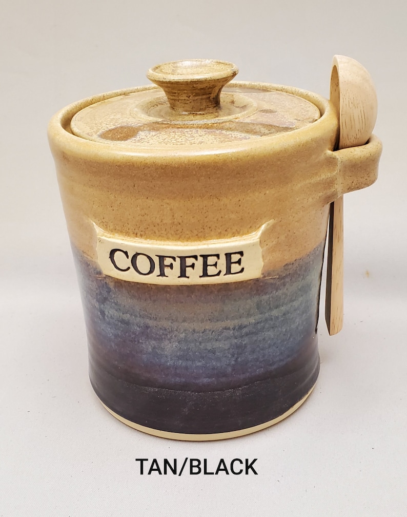 May include: Tan and black ceramic coffee jar with a wooden spoon. The jar has a lid and says "COFFEE" on the front.
