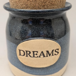 May include: A small, handcrafted ceramic jar with a cork lid. The jar is primarily navy blue with a light blue and cream-colored band. The word "DREAMS" is printed on an oval label on the front.