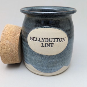 May include: A handmade ceramic jar with a cork lid. The jar is a dark blue with lighter blue and white accents. The words "BELLYBUTTON LINT" are printed on an oval label on the front of the jar. The cork lid is light brown.