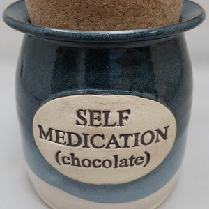 Puede incluir: Tarro de cer&aacute;mica azul y blanco con tapa de corcho. El tarro tiene una etiqueta ovalada blanca con el texto "AUTOMEDICACI&Oacute;N (chocolate)".