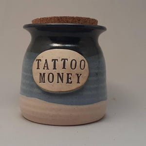 May include: A blue and white ceramic jar with a cork lid. The jar has a white oval label with the text "TATTOO MONEY" in black.
