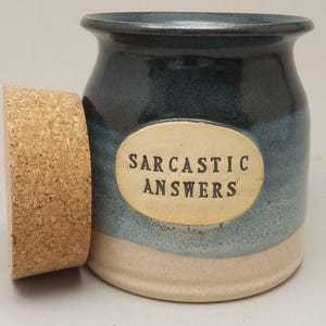 May include: A ceramic jar with a cork lid. The jar is blue and beige with the words "SARCASTIC ANSWERS" on an oval label. The jar is designed to hold small items.
