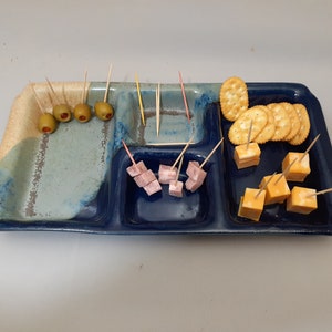 May include: A blue and white ceramic serving tray with four compartments. The tray is filled with snacks, including olives, cheese cubes, ham cubes, and crackers.