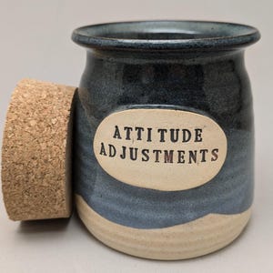 May include: A handmade ceramic jar with a cork lid. The jar is blue and beige with the words "ATTITUDE ADJUSTMENTS" on an oval label. The jar is designed for storage.