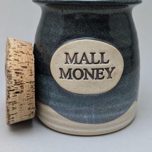 May include: A ceramic jar with a cork lid. The jar is dark blue with a beige base and a beige oval with the words "MALL MONEY" embossed on the front. The cork lid is to the left of the jar.