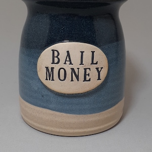 May include: A blue and white ceramic jar with a cork lid. The jar has a white oval label with the text "BAIL MONEY" in black.