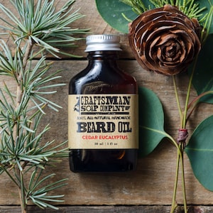 May include: A dark brown glass bottle with a silver lid, labeled "Craftsman Soap Company" and "Beard Oil Cedar Eucalyptus". The bottle is surrounded by green foliage, including pine branches and eucalyptus leaves.