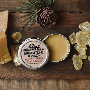 Puede incluir: Un bote de cera para bigote con la inscripción "Craftsman Soap Co. Moustache Wax" abierto, mostrando cera marrón claro. Junto a él, trozos de cera de abeja y resina de pino. Los ingredientes están listados.