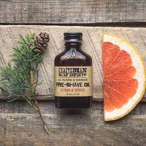 May include: A brown glass bottle with a label that reads "Craftsman Soap Company" and "Pre-Shave Oil Citrus & Spruce". The bottle is sitting on a wooden surface with a small pine branch and a slice of grapefruit.