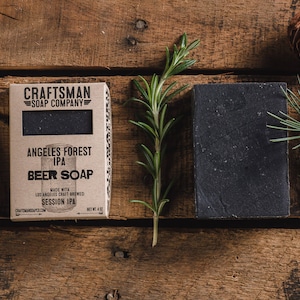 May include: A brown cardboard box with the text "CRAFTSMAN SOAP COMPANY" and "ANGELES FOREST IPA BEER SOAP" printed on it. The box is next to a black bar of soap on a wooden surface.