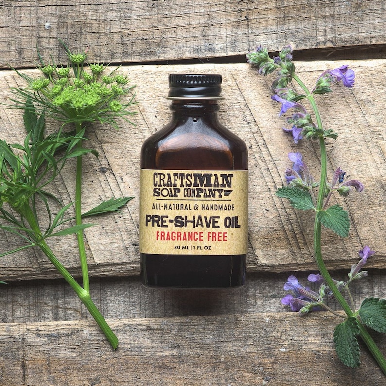 May include: A brown glass bottle with a label that reads "Craftsman Soap Company" and "Pre-Shave Oil Fragrance Free". The bottle is sitting on a wooden surface with green and purple flowers.