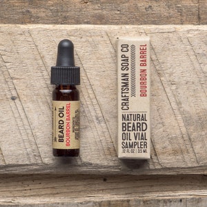 Op de afbeelding: Een klein bruin glazen flesje met een druppelaar, met het label "Beard Oil Bourbon Barrel". Ernaast ligt een bruine kartonnen doos met de tekst "Craftsman Soap Co Bourbon Barrel Natural Beard Oil Vial Sampler 1/2 fl oz | 3.5 ml".