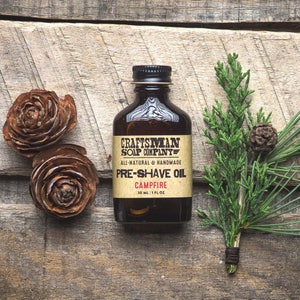 May include: A brown glass bottle with a black label that reads "Craftsman Soap Company" and "Pre-Shave Oil Campfire 30 ml 1 fl oz". The bottle is sitting on a wooden surface with pine needles and pine cones.