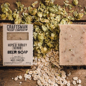 May include: A brown cardboard box with the text "CRAFTSMAN SOAP COMPANY" and "HOPS & BARLEY SCRUB BEER SOAP" printed on it. The box is sitting on a wooden surface with hops and oats scattered around it. The soap bar is visible in the background.