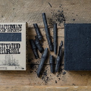 May include: A bar of black activated charcoal soap with a brown label that reads "Craftsman Soap Company" and "Activated Charcoal". The label also includes the text "Net Wt. 4 oz" and "craftsmansoap.com". The soap is surrounded by pieces of charcoal on a wooden surface.
