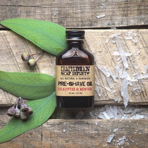 May include: A brown glass bottle with a label that reads "Craftsman Soap Company" and "Pre-Shave Oil Eucalyptus & Menthol". The bottle is sitting on a wooden surface with eucalyptus leaves and clear crystals.