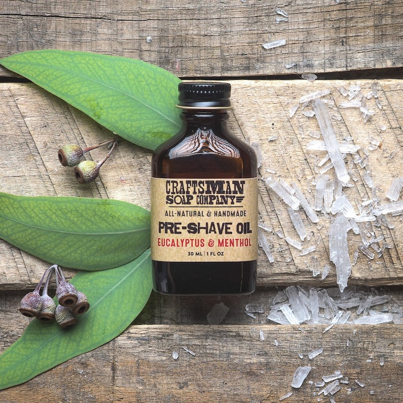 May include: A brown glass bottle with a label that reads "Craftsman Soap Company" and "Pre-Shave Oil Eucalyptus & Menthol". The bottle is sitting on a wooden surface with eucalyptus leaves and clear crystals.