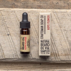 Op de afbeelding: Een klein bruin glazen flesje met een druppelaar, met het label "Beard Oil Hops Flower". Het flesje ligt naast een kartonnen doos met het label "Craftsman Soap Co. Hops Flower Natural Beard Oil Vial Sampler 1.2 fl oz | 35 ml".