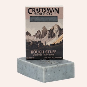 May include: A rectangular bar of grey soap with dark speckles sits in front of its cardboard packaging. The box features a mountain landscape illustration and the text CRAFTSMAN SOAP CO, ROUGH STUFF, UNSCENTED, HEAVY SCRUB, NET WT 113 GRAMS.