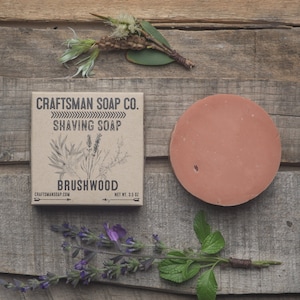 Puede incluir: Una caja de cartón marrón con el texto "Craftsman Soap Co. Shaving Soap Brushwood" y "Net Wt. 3.5 oz" en ella. Un disco redondo de jabón de afeitar de color naranja claro está al lado de la caja sobre una superficie de madera.