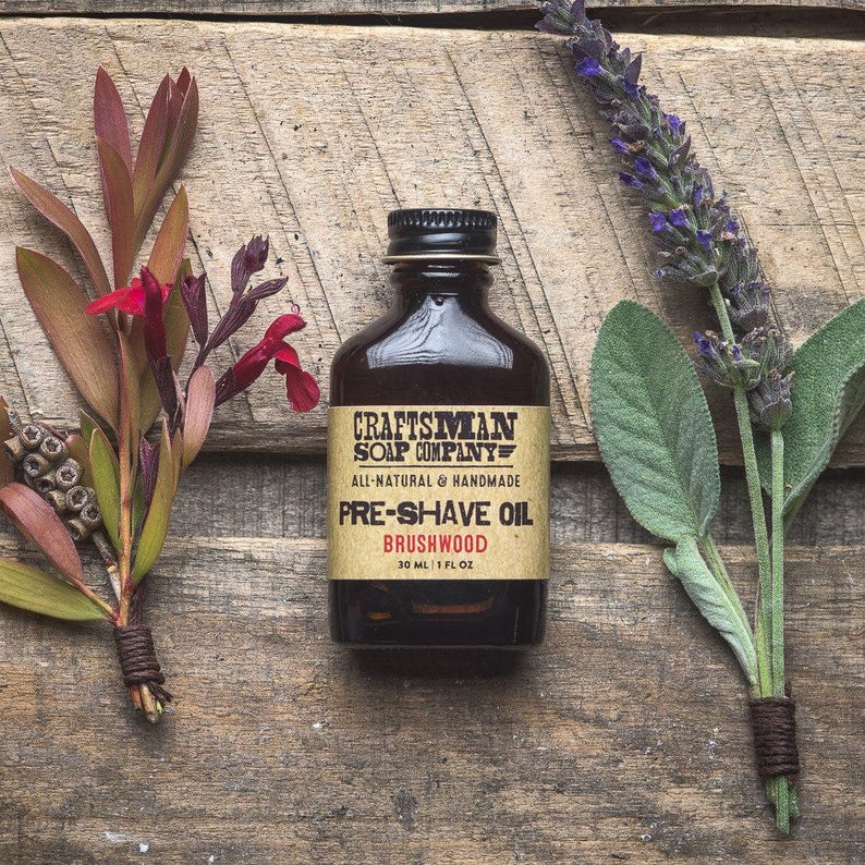 May include: A brown glass bottle with a label that reads "Craftsman Soap Company" and "Pre-Shave Oil Brushwood". The bottle is on a wooden surface with green and purple flowers.