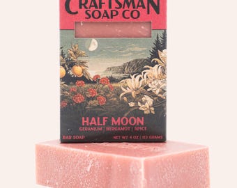 Op de afbeelding: Een rechthoekig stuk roze zeep voor de verpakking. De doos bevat de tekst CRAFTSMAN SOAP CO, HALF MOON, GERANIUM, BERGAMOT, SPICE en NET WT 113 GRAMS, met een illustratie van een maanverlicht landschap, bloemen en vruchten.