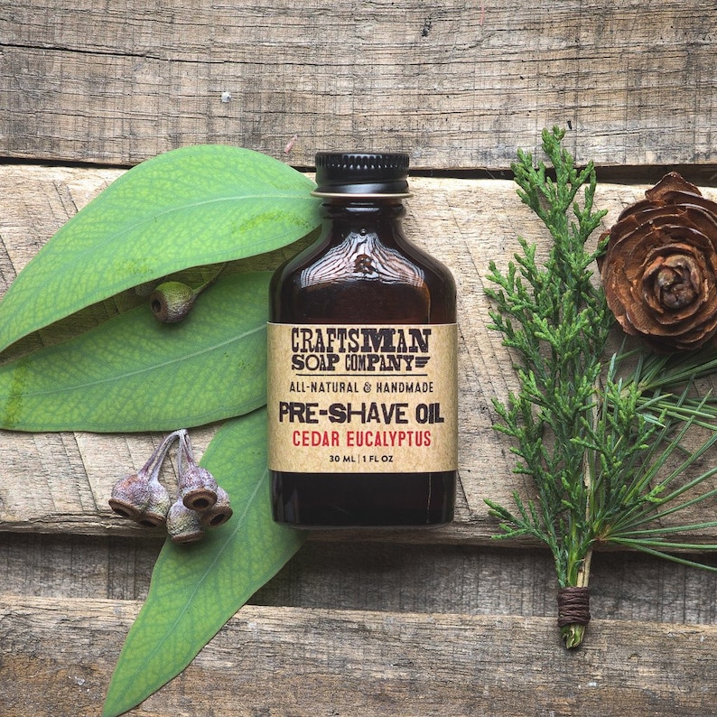 May include: A brown glass bottle with a black label that reads "Craftsman Soap Company" and "Pre-Shave Oil Cedar Eucalyptus". The bottle is sitting on a wooden surface with green leaves and a brown pine cone.