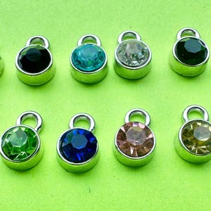 May include: A collection of twelve small, round charms with silver-colored metal settings. Each charm features a different colored gemstone, including red, black, teal, clear, green, blue, pink, and yellow. The charms have a loop at the top for attaching to jewelry.