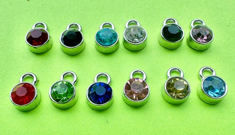 May include: A collection of twelve small, round charms with silver-colored metal settings. Each charm features a different colored gemstone, including red, black, teal, clear, green, blue, pink, and yellow. The charms have a loop at the top for attaching to jewelry.