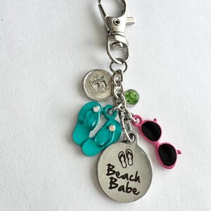 May include: Silver-tone keychain with beach-themed charms. Includes turquoise flip-flops, pink sunglasses, a round charm with "Beach Babe" text, a green gem, and a coin-shaped charm. The keychain has a lobster clasp.