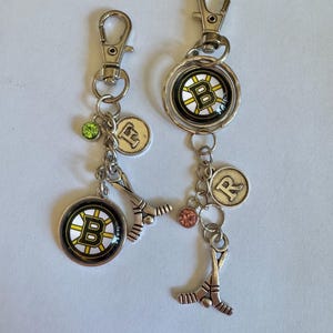 May include: Two silver keychains with lobster claw clasps. Each keychain features a Boston Bruins logo charm, hockey stick charm, and a letter charm. One has a green gem, the other an orange gem.