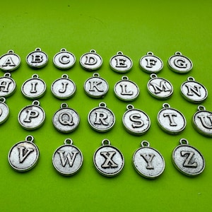 May include: Silver-colored alphabet charms, each with a different letter from A to Z, are displayed against a bright green background. The charms are round with a loop at the top for attaching to jewelry or other crafts.
