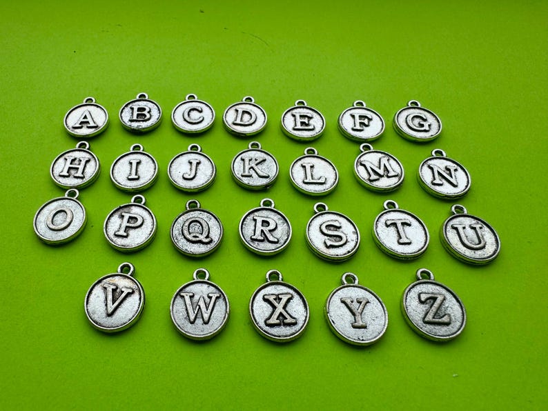 May include: Silver-colored alphabet charms, each with a different letter from A to Z, are displayed against a bright green background. The charms are round with a loop at the top for attaching to jewelry or other crafts.