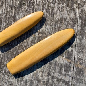 May include: Two wooden paddles, one slightly larger than the other, are lying on a weathered wooden surface. The paddles are a light brown color and have a smooth, polished finish.