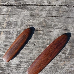 May include: Two wooden paddles, one is a darker brown and the other is a lighter brown, both have a wood grain pattern. The paddles are laying on a wooden surface.