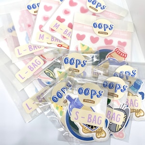 Puede incluir: Surtido de bolsas de plástico transparente llenas de pegatinas coloridas. Las pegatinas presentan varios diseños, incluidos corazones, peces y una botella azul. Las bolsas están etiquetadas con el texto "L-Bag", "M-Bag", "S-Bag" y "Oops Stickers".