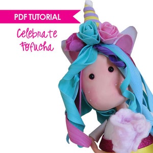 May include: A blue and pink unicorn doll with a rainbow horn and a pink and blue flower crown. The doll has a white dress and a pink and white fluffy tail. The doll is holding a pink and white fluffy cloud. The text "Celebrate Tofiicha" is written in pink letters on a pink banner.