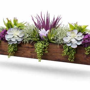 May include: A rectangular dark brown wooden planter box filled with an assortment of artificial succulents and greenery. The arrangement features light green, pale blue, and deep purple plants, including spiky leaves and trailing vines.