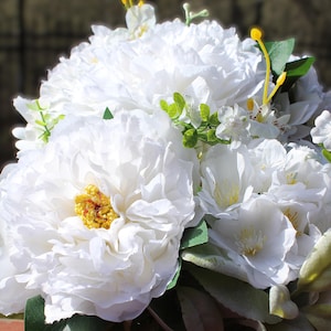 May include: A close-up of a bouquet of white flowers, featuring large, ruffled petals with yellow centers. Green leaves and stems provide contrast. The arrangement includes smaller white blossoms and yellow accents.