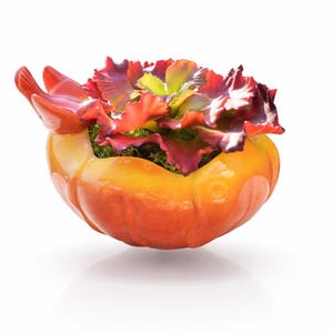 May include: A ceramic planter in the shape of a pumpkin, with a glossy orange and yellow finish. The planter is filled with a colorful arrangement of artificial flowers in shades of red, purple, and green.