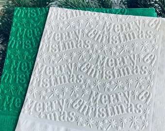 May include: White and green embossed paper napkins with the words "Merry Christmas" and snowflake designs. The napkins are stacked, with the white napkin on top and a green napkin underneath. Festive holiday decor.