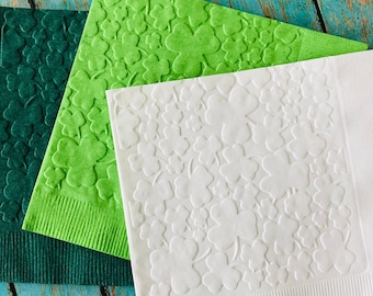 May include: Three paper napkins in shades of green and white, each with a clover pattern. The napkins are arranged on a weathered, turquoise-colored wooden surface. The napkins are a festive decoration for St. Patrick's Day.